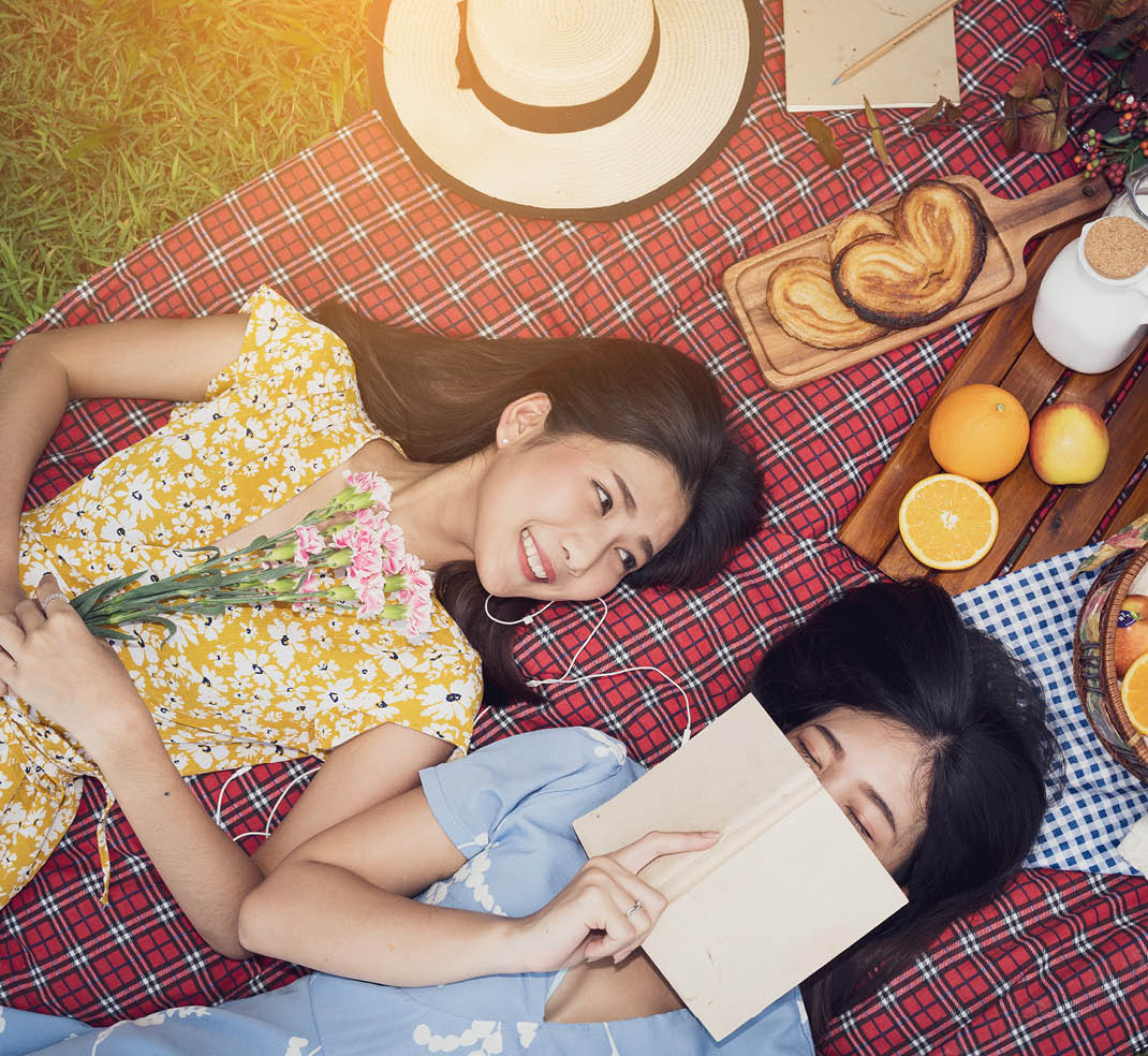 Couple of girls going picnic in a park, reading a book together and feeling friendly.