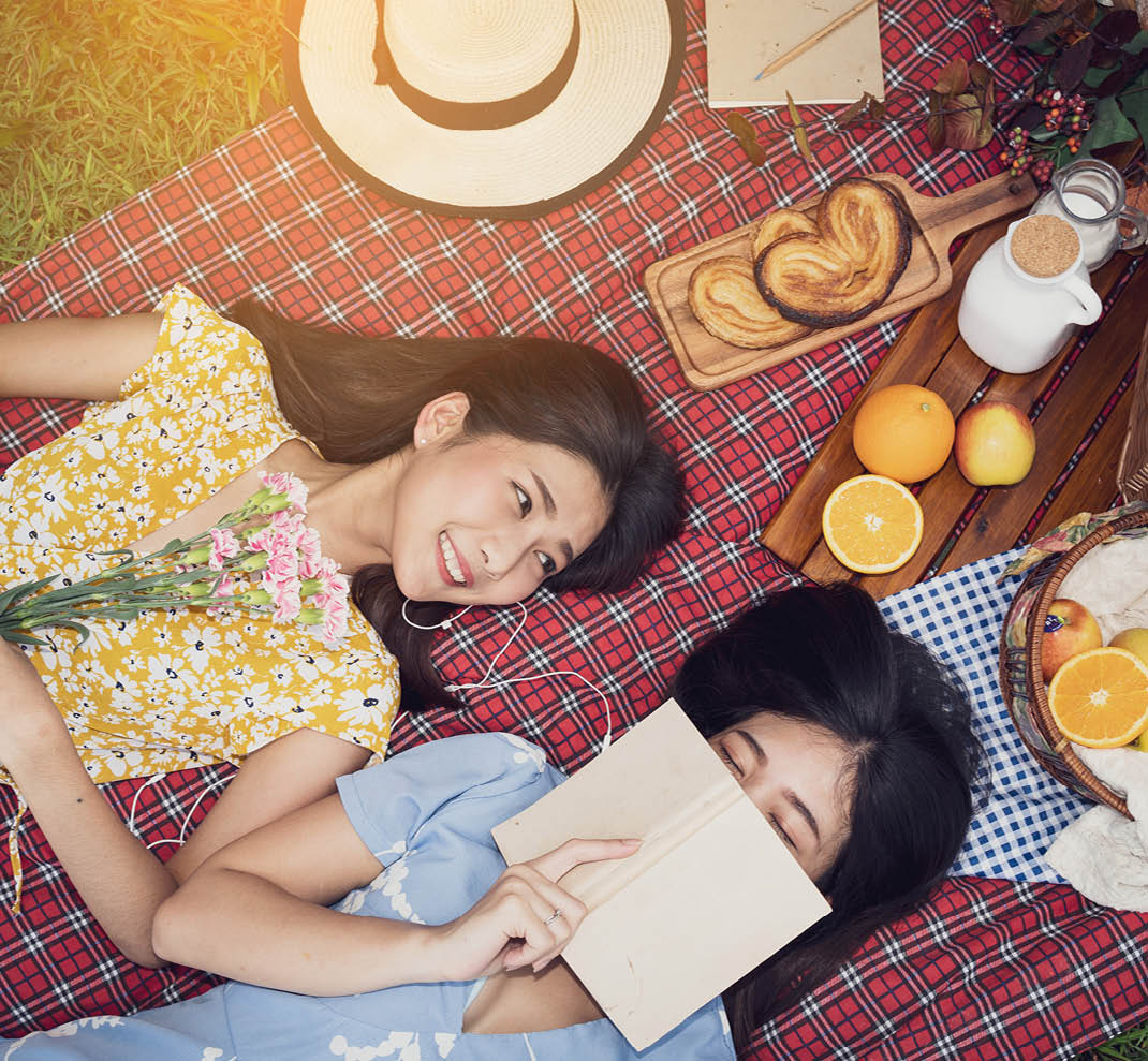 Couple of girls going picnic in a park, reading a book together and feeling friendly.